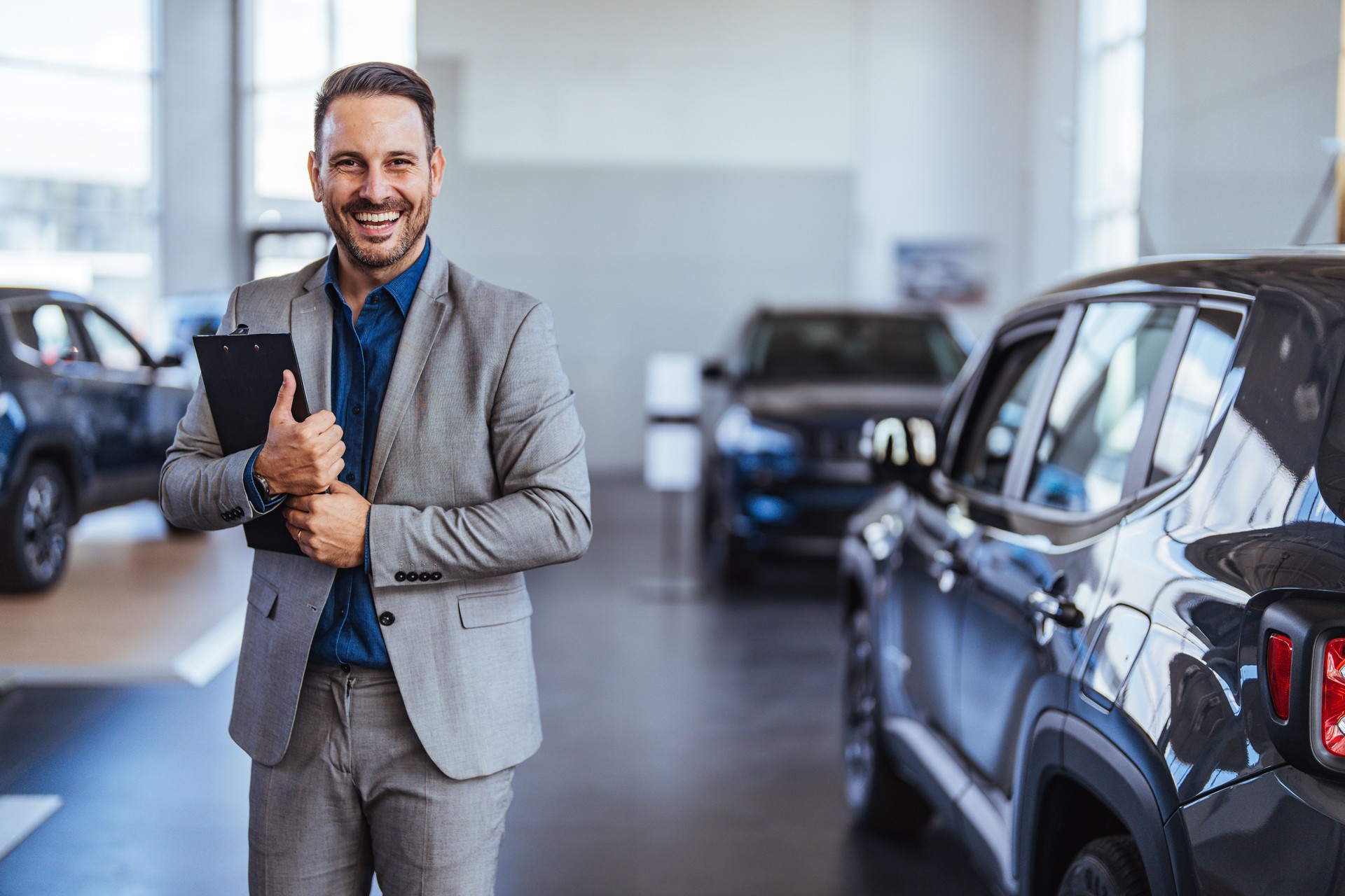 Happy salesperson standing in a car showroom looking at camera.