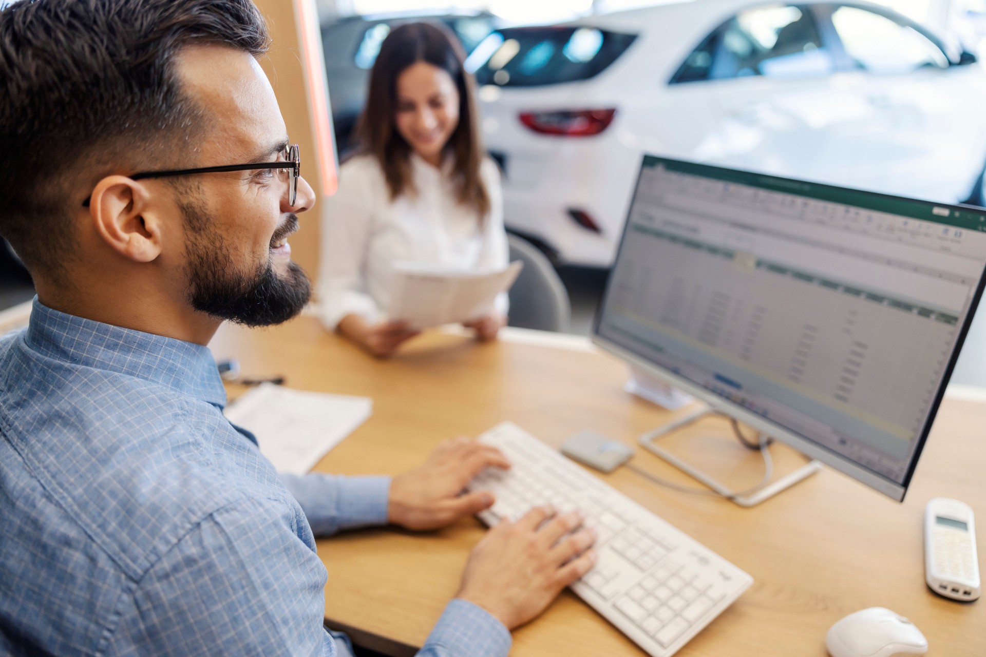 Smiling car dealer sitting at car salon and typing documents on computer while buyer reading contract